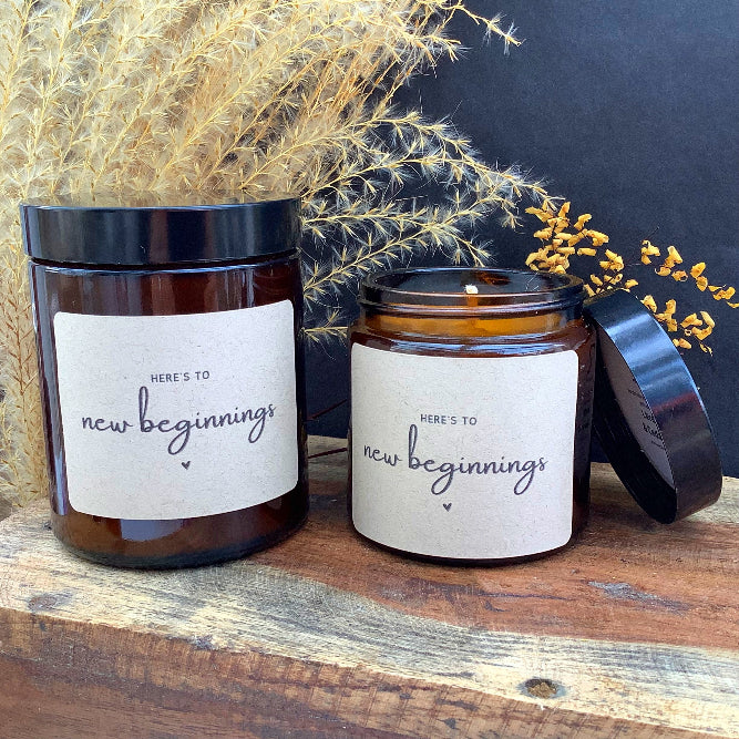 Two candles with 'Here's to new beginnings' labels on a wooden surface with dried plants.