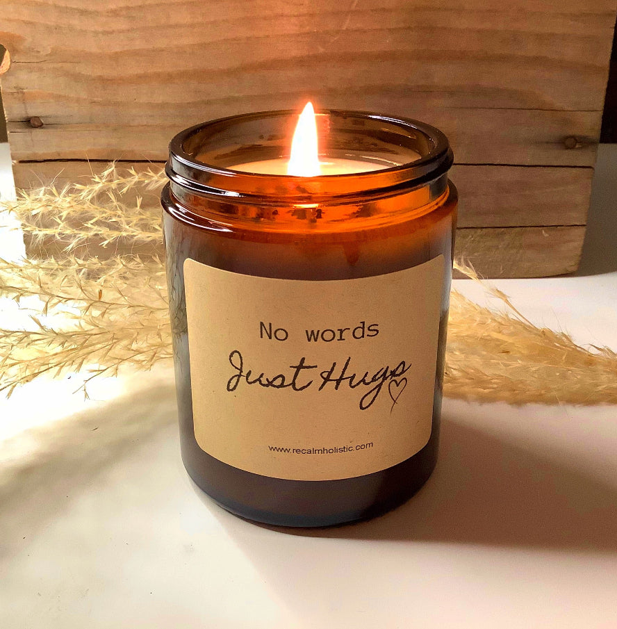 Candle in an amber jar with a label reading 'No words just hug' on a wooden surface.