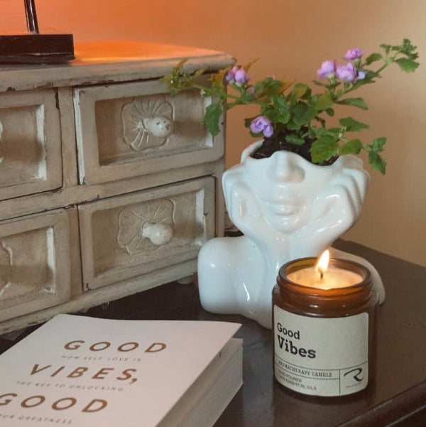 Candle with 'Good Vibes' label on a table next to a book and decorative lamp