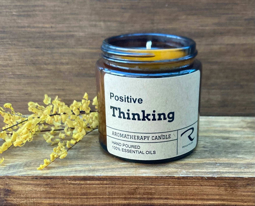 Aromatherapy candle labeled 'Positive Thinking' on a wooden surface with yellow flowers.