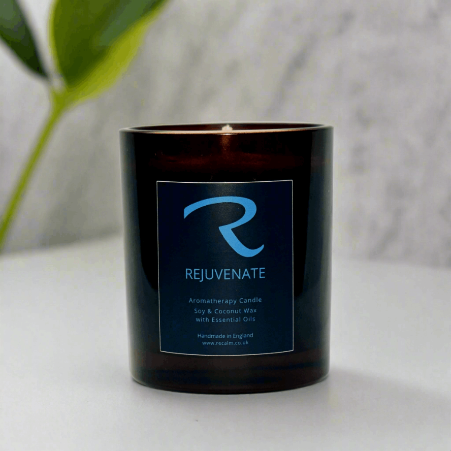 Black candle with 'Rejuvenate' label on a white surface with a blurred green plant in the background