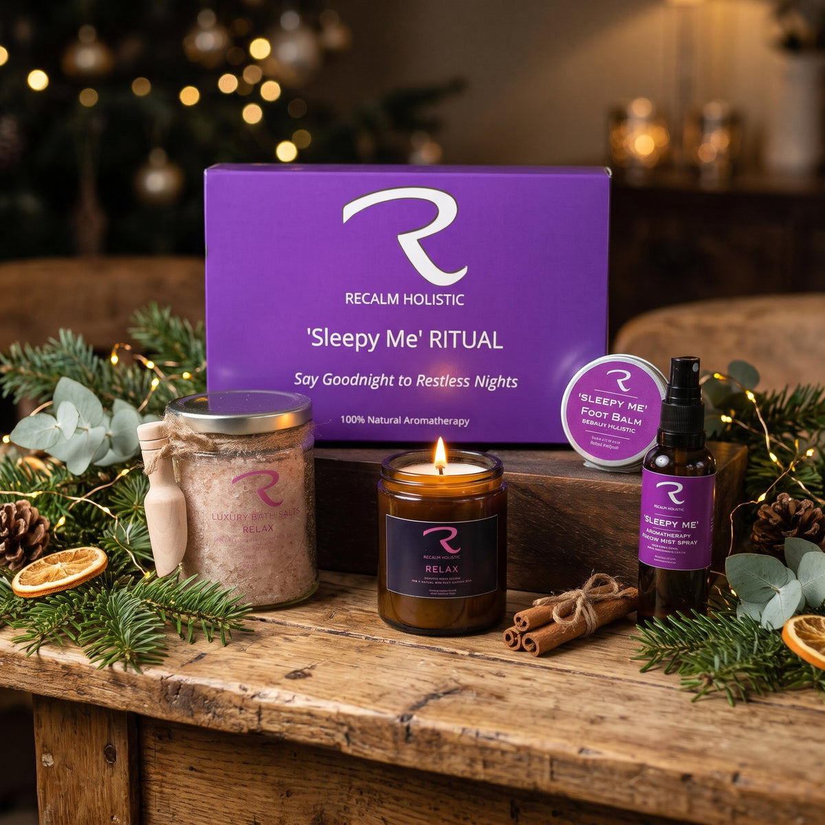 A cozy holiday gift set on a rustic wooden table, featuring a purple box labeled 'Recalmb Holistic Sleepy Me Ritual', a lit amber candle, a jar of bath salts, a small bottle of essential oil, and a round tin of foot balm, surrounded by eucalyptus, pine branches, cinnamon sticks, and warm bokeh lights in the background.