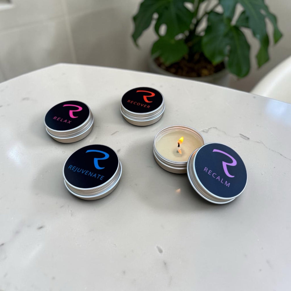 Four small round silver candle tins on a white marble surface, with one candle open and lit. The lids have dark blue labels with a stylized 'R' logo and words including RELAX, RECOVER, REQUENATE, and RECALM, finest aromatherapy candles in the UK and the world