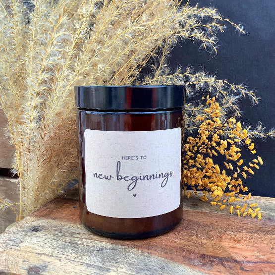 Candle jar with 'Here's to new beginnings' label on a wooden surface with decorative plants.