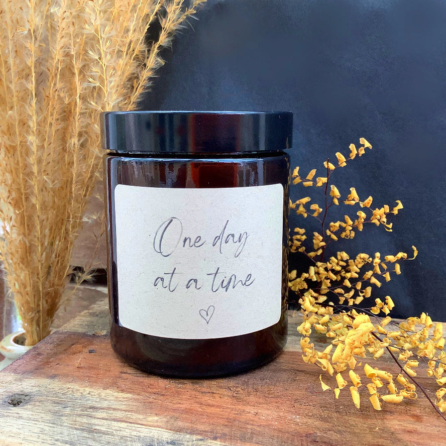 Brown candle jar with 'One day at a time' label on a wooden surface with decorative plants.