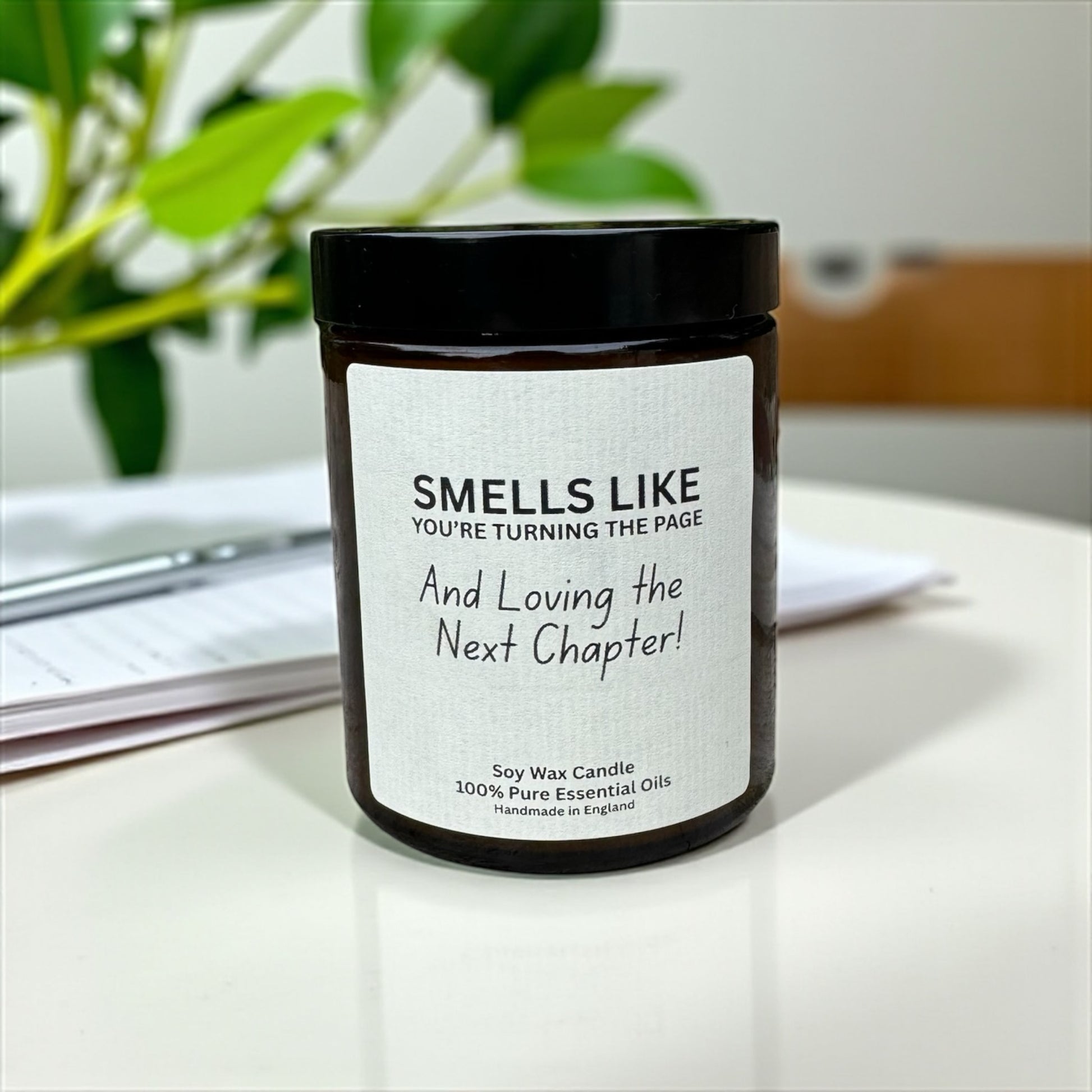 Candle jar with a humorous label on a desk with a plant in the background, Smells like you're turning the page and loving the next chapter