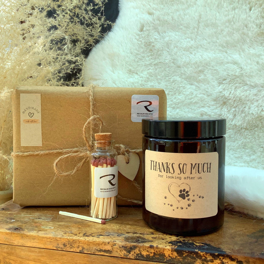 Candle with 'Thanks so much' label, small bottle with matches, and wrapped gift on wooden surface by ReCalm Holistic