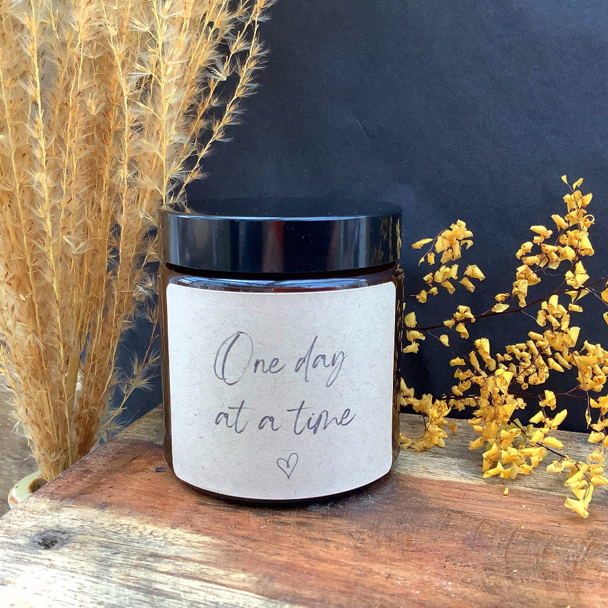 Jar with 'One day at a time' text on a wooden surface with decorative plants.